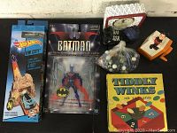 Full view of all toys displayed on a black surface including the Hot Wheels, Batman figure, marbles, Tiddly Winks box, pencil sharpener, and clock