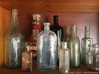 Nine vintage and antique glass bottles displayed on a wooden shelf, showing varying colors and sizes including clear, green, amber, and black.