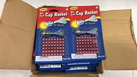 Box containing multiple Toysmith Die Cast Cap Rocket packages, each with 48 blast caps and a metallic silver cap rocket toy.