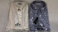 Lot of two new men's dress shirts in plastic packaging, one white and one blue, showing front collar and tags.