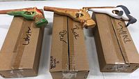 Photo showing three wooden rubber band shooters shaped like a lobster, deer, and dolphin, placed on cardboard boxes labeled accordingly.