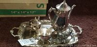 Full view of silver plated tea set with tray, teapot, cream and sugar dishes, and two bird figurines on dark carpet.