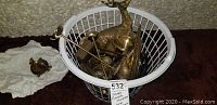 Photo showing a white basket filled with various brass items including serving platters, wildlife figurines, and candle holders, along with a small bird figurine outside the basket.