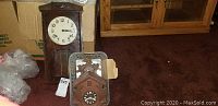 Image showing rectangular wooden wall clock with white clock face and Arabic numerals, alongside two wooden cuckoo clock cases in plastic bin on carpeted floor