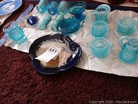 Overview of all blue glass items including candle holders, creamers, sugar dishes, salt shaker and display plate on white paper on carpet.