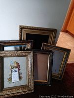 Five antique picture frames including one with a signed needlepoint artwork visible in front with four additional empty frames behind leaning against wall.