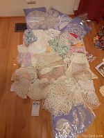 Over twenty crocheted doilies piled on wooden floor showing variety of sizes and styles including some packaged and some loose.