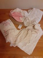 Photo showing a group of three table cloths, pink crocheted edge cloth, and two white embroidered cloths, bundled on a wooden surface