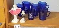 Six blue Shirley Temple glasses with picture and signature, two salt bisque dancing figurines on wooden shelf.