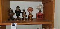 Group of nine vintage oil lamps on wooden shelf, including fireplace-style lamp on right with red base and glass chimney.