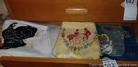 Three decorative linens displayed in a drawer, showing rooster, bird, floral, and winter scenes