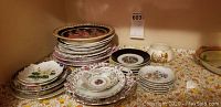 Multiple stacks of decorative plates and small bowls with various floral and scenic patterns, shown on a linen surface.
