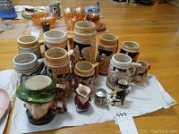 Group photo of eleven beer steins, three creamers, and one beer glass arranged on white cloth on a wooden table showing various designs and sizes