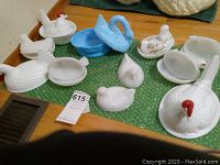 Overview of the collection showing ten white hen-shaped milk glass dishes and one blue swan dish on green bubble wrap.
