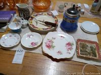 Five dinner plates, four tea saucers, one tea decanter, two creamers, one sugar bowl, one teacup, one meat fork all arranged on table with various patterns and colors primarily floral and blue and gold decanter.