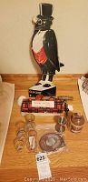 Full lot view showing the Old Crow advertising figurine, shot and whiskey glasses in tartan box, antique corkscrew, and two small pitchers on wooden surface.