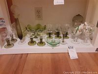 Collection of green glass items including ten goblets, St. Patrick's Day stemware, vase, candy dishes, and decanter arranged on shelf