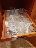Top view of drawer containing mini punch bowl, small cups, covered candy dish, candle holders, and wine bottle stoppers all in clear glass.