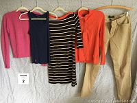 Five Ralph Lauren women's clothing items hanging on a wall: pink wool sweater, navy cotton tank top, brown and white striped knit dress, orange cotton sweater, tan cotton lycra jeans.