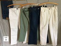 Front view of 5 pairs of designer jeans in various colors hung on wooden hangers.