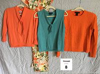 Front view of three cashmere sweaters (two orange, one aqua) and a large floral silk scarf underneath.
