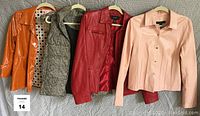 Four outerwear items displayed on hangers on a grey background, showing front view: an orange vinyl jacket, black and white check vest with hood, red leather jacket, and pink leather jacket.