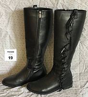 Front side view of the pair of black leather tall boots with fringe and side zipper.