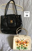 Front view of a large black leather tote bag with flap closure and a vintage white handbag with a butterfly design, both shown on a hanger and flat on a bedspread.