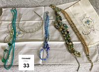 Full view of 2 beaded necklaces in blue and turquoise, rhinestone headband, faux pearl necklace, vintage costume necklace with green and gold beads with some paint peeling, all arranged on embroidered fingertip towels.