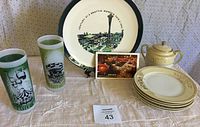 Photo of two green Seattle tumbler glasses, sugar bowl, six china plates, commemorative World's Fair plate, and folded postcard string all displayed on white cloth.