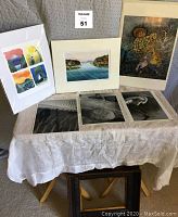 Lot of seven art prints displayed on a table with lace tablecloth including colorful prints and three black and white wildlife photos, some matted and some loose.