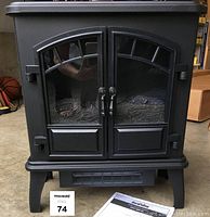Front view of electric heater showing black metal body with glass front doors and faux flame inside. Instruction booklet visible in foreground.