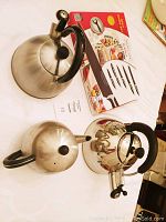 Three stainless steel teapots with black handles arranged on a surface alongside the Royalty Line 10-piece knife set box with knives depicted on the packaging.