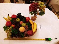Wide view of oval basket with assorted faux fruit and the glass candle holder with faux grapes on a table, including measuring tape showing basket size