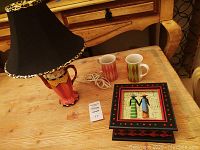 Photo showing teapot lamp with black leopard print lampshade, two tea mugs, and decorative tea box on wooden table