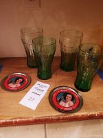 Four Coca Cola drinking glasses and two metal Coca Cola coasters on a wooden surface, clearly showing variation in glass colors and coaster design.
