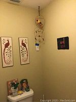 Parrot decor arranged on a wall including two rectangular tile plaques with maroon parrot designs, a slate parrot painting, and smaller items placed on toilet tank.