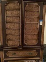 Full view of the wooden armoire with two doors and a drawer below, showing the woven wicker paneling and wooden frame.