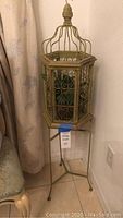 Full view of wooden bird cage on tall metal stand placed against a corner wall with drapes and chair nearby.