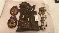 Full lot contents showing carved wood Egyptian lady plaque, bird wall decor, and metal wind chime on white fabric surface