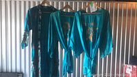 Three teal blue silk robes hanging on wooden hangers. They have gold embroidery designs on the front and matching belts.