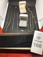 Photo showing 4 gold-colored necklaces, 2 jewelry boxes (one with pearl pendant, one with clear stones), and small gold fish charm on black mat