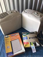 Two beige Hunter Quiet Flo air purifiers sitting side by side along with their replacement filters on a blue surface.