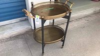 Full view of the brass tray cart showing two oval brass trays supported on a black foldable metal stand with caster wheels.