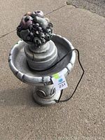 Full view of the heavy grey birdbath on concrete with black cable visible and a lot tag attached.