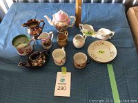 Thirteen assorted tea cups, saucers, teapots, creamers, and mugs arranged on blue cloth surface from a side angle.