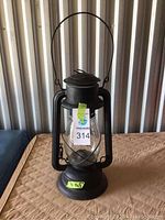 Full front view of black metal railway lantern on brown quilted surface. Wire handle extended. Number label taped to glass globe.