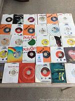 Photo showing all 30 vinyl 45 RPM records laid out on a table in various sleeves with visible labels and some album cover art.