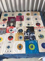Top-down image showing 30 vinyl 45 rpm records mostly in paper sleeves laid out on flat surface.