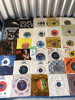 Lot of 30 45 RPM vinyl records arranged in rows, showing varied record label colors and paper sleeves with different branding and logos